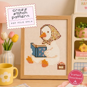 May include: A cross stitch pattern PDF file featuring a white duck wearing a colorful patterned headscarf while reading a blue book with pink hearts. The duck has orange feet and a beak, sitting next to a small brown basket on a light fabric background.