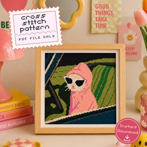 May include: A framed cross stitch pattern featuring a cat wearing a pink hoodie and black sunglasses while sitting in a car. The text on the pattern reads cross stitch pattern PDF FILE ONLY. The design uses green, black, and pink thread colors.