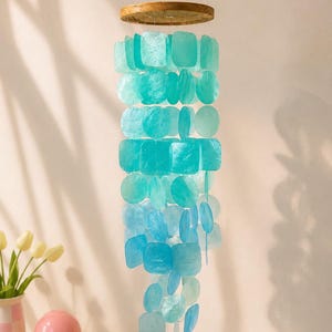 Può includere: Una giostrina sospesa con una parte superiore circolare in legno e molteplici fili di dischi di conchiglia traslucidi e iridescenti in tonalità turchese, acqua e blu, disposti in un motivo a cascata che sfuma dal chiaro allo scuro.