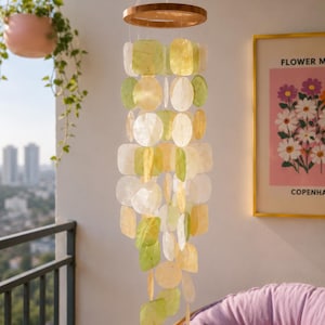 May include: A hanging mobile featuring a circular wooden frame with multiple strands of translucent, flat, circular shell discs in shades of pale yellow, light green, and white, suspended by thin strings to create a layered, cascading decorative display.