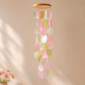 Op de afbeelding: Een hangende mobiel met meerdere doorschijnende, platte, ronde en vierkante schelpstukjes in roze, lichtgroene en witte tinten. De stukjes hangen aan een ronde houten bovenkant met behulp van dunne, doorzichtige koordjes in een trapsgewijze opstelling.