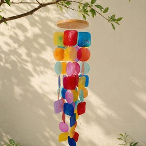 May include: A hanging wind chime featuring a circular wooden top piece with multiple strands of translucent, colourful capiz shell discs and squares in shades of red, orange, yellow, blue, and purple, suspended by thin clear nylon strings.