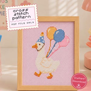 May include: A cross stitch pattern PDF file featuring a white goose wearing a blue party hat with white dots, holding three balloons in yellow, blue, and pink. The design is set against a light pink background and displayed in a light wood frame.
