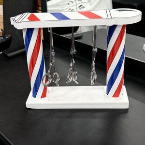 May include: A barber pole-themed stand featuring two vertical pillars with red, white, and blue diagonal stripes, connected by a horizontal top bar and a white base. Three silver metal hair-cutting shears hang vertically from the top bar.