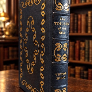 May include: A dark blue hardback book titled The Toilers of the Sea by Victor Hugo. The cover and spine feature intricate gold foil scrollwork patterns. The spine includes raised bands and clear gold lettering displaying the title and author name.
