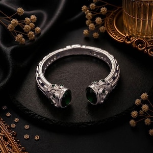 Può includere: Un bracciale a fascia in argento con una banda testurizzata a motivi geometrici ripetuti e due pietre ovali verde scuro alle estremità aperte, arricchito da piccole pietre circolari verdi incastonate lungo la struttura in metallo.