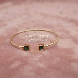 Könnte beinhalten: Ein goldfarbenes offenes Armband mit strukturiertem Band und zwei quadratischen grünen Steinen an den Enden. Das Armband hat eine Länge von 5,51 cm und eine Breite von 5,51 cm, wie durch den Text direkt auf dem Bild angegeben.