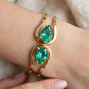 May include: A gold-toned open cuff bracelet featuring two teardrop-shaped, faceted emerald green stones. The metal band curves around each stone, creating a symmetrical design that leaves an open space between the two ends of the bracelet.