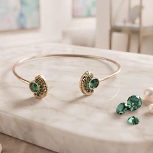 May include: A gold-toned open cuff bracelet featuring two semi-circular ends, each set with a central round green gemstone surrounded by smaller green stones. Three loose, faceted green gemstones and two white pearls lie nearby on the surface.