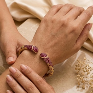 May include: A gold cuff bracelet featuring a textured surface and two rounded ends encrusted with small, vibrant red stones. The bracelet has a wide, open design that wraps around the wrist, showcasing a detailed pattern of red stones set against a gold-toned metal base.