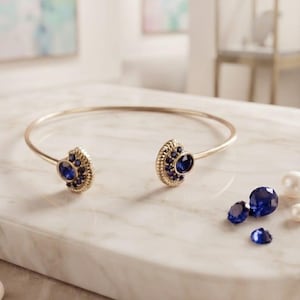 May include: A gold-toned open cuff bracelet featuring two semi-circular ends, each set with a central round blue stone surrounded by smaller blue accent stones. The bracelet is displayed alongside several loose blue faceted stones and white pearls.