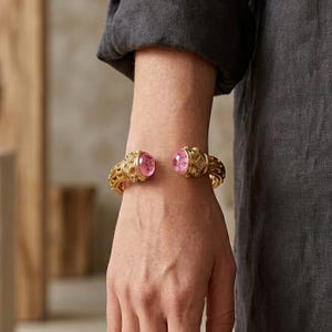 May include: A gold cuff bracelet featuring an open design with two rounded, polished pink gemstones at each end. The metal band displays an intricate, textured pattern across its surface, creating a detailed appearance on the wrist.