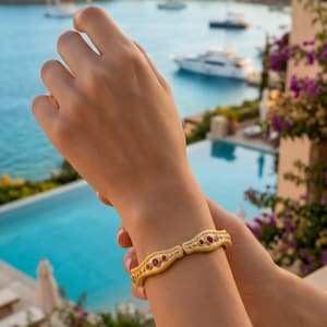 May include: A gold-toned hinged bangle bracelet featuring a textured surface and inlaid with small, round, deep pink gemstones. The bracelet has a slightly curved, organic shape and is secured with a visible hinge mechanism on the side.