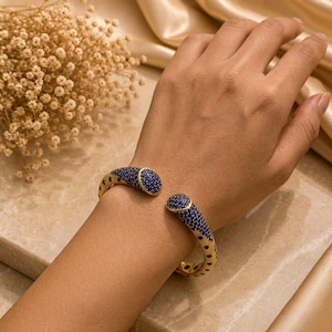 Puede incluir: Un brazalete tipo puño dorado con una superficie texturizada y un patrón denso de pequeñas piedras azul oscuro. El brazalete tiene un diseño abierto con dos extremos redondeados incrustados de piedras que se enfrentan entre sí.