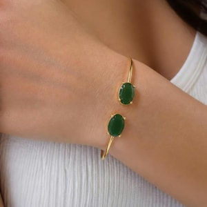 Op de afbeelding: Een open gouden armband met twee ovaalvormige donkergroene stenen die aan beide uiteinden in gouden klauwen zijn gezet. De armband heeft een dunne, gepolijste gouden band die om de pols buigt en de twee gladde, diepgroene cabochon-edelstenen toont.