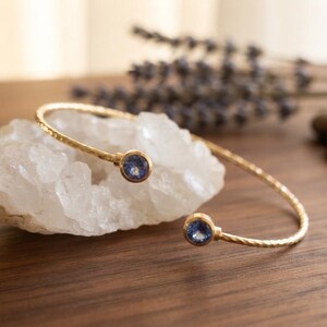 Puede incluir: Una pulsera abierta de tono dorado con una banda de metal martillado y texturizado. Los dos extremos de la pulsera terminan en cristales azules redondos y facetados que aportan un brillo sutil al diseño general de esta pieza de joyería.