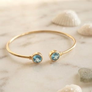 Könnte beinhalten: Ein goldfarbenes, offenes Armband mit zwei runden, hellblauen facettierten Steinen an den Enden. Der Metallreif ist dünn und poliert und bildet eine Kreisform mit einer Lücke zwischen den beiden Edelsteinfassungen.