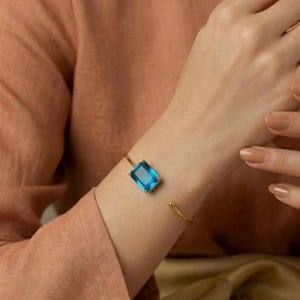 以下が含まれることがあります： 4本の爪で留められた長方形のファセットカットされた青い宝石が特徴の、ゴールドトーンのオープンカフブレスレット。ブレスレットは細い磨かれた金属バンドでできており、開いた両端には小さな丸いゴールドトーンの球体がついています。