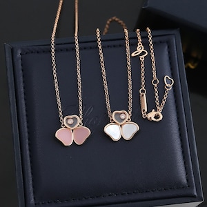 May include: Two rose gold necklaces featuring a three-leaf clover design. Each pendant includes a small heart-shaped element with a floating diamond and two petals, one with pink stone inlay and one with white mother-of-pearl inlay, on a chain.