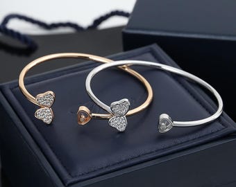 May include: Two open-ended cuff bracelets, one in rose gold and one in silver-toned metal. Each bracelet features a heart-shaped motif encrusted with small clear stones and a smaller heart-shaped charm containing a single floating clear stone.
