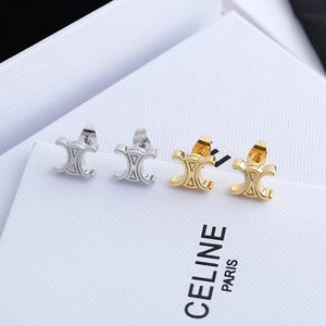 May include: Two pairs of stud earrings, one silver-toned and one gold-toned, each featuring a symmetrical triomphe-inspired logo design. The earrings are positioned on a white textured surface with the black printed text CELINE PARIS visible.