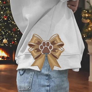 May include: A white sweatshirt featuring a large, centred graphic of a tan and brown ribbon bow with a central paw print design. The sweatshirt is worn over light blue denim jeans, showcasing the detailed illustration of the bow and paw.