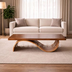 Op de afbeelding: Een moderne salontafel van massief hout met een unieke, gebogen basis. Het tafelblad is een rijk, donkerbruin en de basis heeft een lichtere houttint. De tafel staat in een woonkamer met een cr&egrave;mekleurige bank en een neutraal tapijt.