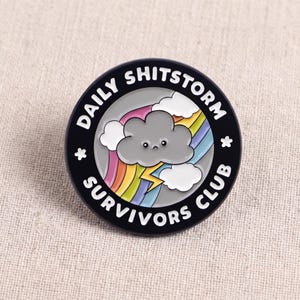Może przedstawiać: Okrągła emaliowana przypinka z czarną obwódką i białym napisem DAILY SHITSTORM SURVIVORS CLUB. W centrum znajduje się smutna szara chmura z twarzą, żółta błyskawica, kolorowa tęcza i białe chmury na szarym tle.