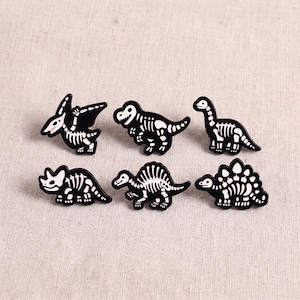 May include: A set of six enamel pins featuring black dinosaur silhouettes with white skeletal details. The collection includes a pterodactyl, a tyrannosaurus rex, a long-necked sauropod, a triceratops, a spinosaurus, and a stegosaurus design.