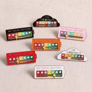 Puede incluir: Una colección de siete pines de esmalte con diseños rectangulares en forma de batería y de nube. Cada pin muestra el texto MY SOCIAL BATTERY o MOOD TRACKER, un indicador deslizante y una serie de cuadrados de colores con expresiones faciales.