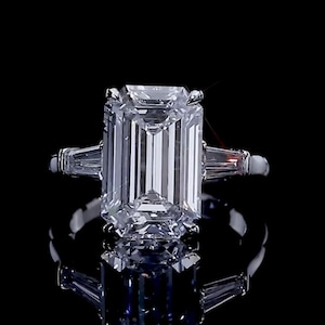 Puede incluir: Un anillo de tono plateado con una gran piedra central rectangular de corte esmeralda con apariencia clara y facetada. La piedra central está flanqueada por dos piedras más pequeñas de corte baguette cónico a cada lado, sobre una banda de metal pulido.