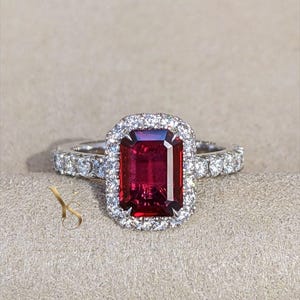 Könnte beinhalten: Ein Ring mit einem großen, rechteckigen, tiefroten Edelstein in einer silberfarbenen Metallschiene. Der Mittelstein ist von einem Kranz aus kleinen, runden, klaren weißen Steinen umgeben, wobei weitere weiße Steine in die Ringschiene eingelassen sind.