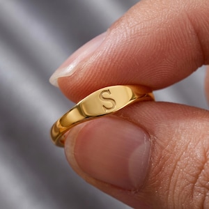 May include: A gold-toned signet ring featuring a smooth, polished surface with the capital letter S engraved in the center. The ring has a rounded band and a flat, oval-shaped face that displays the letter clearly against the metallic finish.