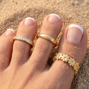 May include: Three gold-toned toe rings worn on three toes. One ring features a row of small clear stones, the second is a plain polished gold band, and the third is a band decorated with a repeating pattern of small gold daisy-shaped flowers.