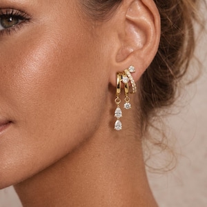 May include: A gold-toned triple hoop earring set featuring two dangling teardrop-shaped clear stones and two round clear stones. The design includes a small hoop with a pavé-style band of clear stones and two plain gold hoops with hanging charms.