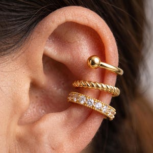 May include: A set of three gold-toned ear cuffs worn on the cartilage. The top cuff is a smooth band with a spherical end, the middle cuff features a twisted rope texture, and the bottom cuff is a band encrusted with clear, round-cut stones.