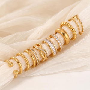 May include: A collection of gold-toned rings and ear cuffs featuring various designs, including a smooth band, a twisted rope texture, a beaded band, an infinity symbol with clear stones, a baguette-cut stone band, and a delicate chain link design.