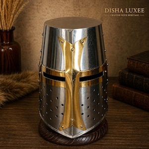 Puede incluir: Un casco de caballero de metal plateado con una cruz dorada superpuesta en la parte frontal, una ranura horizontal para los ojos y múltiples orificios de ventilación. El casco está montado sobre una base de madera circular y muestra el texto DISHA LUXEE.