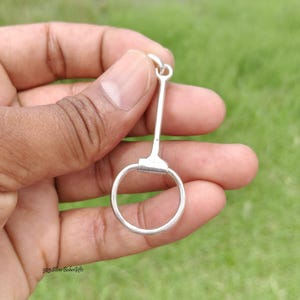 May include: A silver-coloured metal pendant shaped like a horse bit, featuring a circular ring attached to a straight vertical bar with a small loop at the top for suspension. The piece has a smooth, polished finish and is held against a green background.
