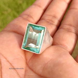 May include: A silver ring featuring a large, rectangular, light blue gemstone. The ring has a simple, modern design with a wide band. The gemstone is set in a bezel setting, and the ring is shown on a hand.