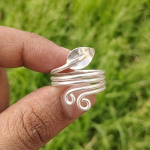 May include: A silver-colored metal ring featuring a wrap-around design. The band consists of multiple coiled wires that terminate in a leaf-shaped element at one end and two small, circular spiral loops at the other end.