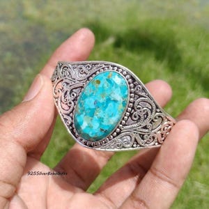 May include: A silver cuff bracelet featuring an oval-shaped turquoise stone with blue and green mottled patterns. The metal band displays intricate, raised scrollwork and filigree detailing surrounding the central gemstone setting.