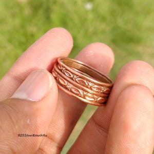 Puede incluir: Primer plano de tres anillos color cobre con un patrón ovalado en relieve. Los anillos están apilados, mostrando el diseño en las bandas exteriores. Se sostienen en una mano sobre un fondo verde borroso. El texto "925SilverBohoGifts" es visible.