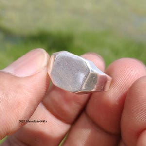 May include: A close-up of a silver signet ring with a geometric, rectangular face. The ring is held between fingers against a blurred green background. The text "925SilverBohoGifts" is visible in the lower left corner.