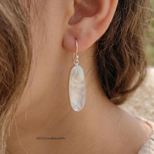 May include: An oval-shaped earring featuring a smooth, iridescent mother-of-pearl inlay set within a polished silver frame. The earring hangs from a simple silver hook wire, displaying a reflective, pearlescent surface with subtle white tones.