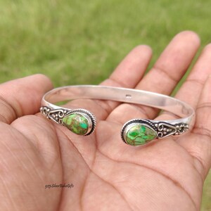 May include: A silver cuff bracelet featuring two teardrop-shaped green turquoise stones at each open end. The metal settings around the stones display intricate twisted rope and beaded detailing. The band is smooth and polished silver.