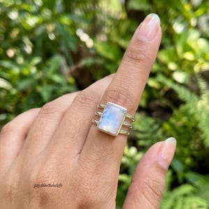May include: A silver ring featuring a rectangular, iridescent moonstone cabochon set in a bezel. The band consists of three parallel silver wires that converge at the back. The text 925 Silver BohoGifts is visible on the lower left side.