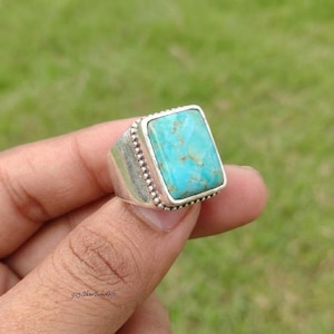 May include: A silver ring featuring a rectangular turquoise stone with brown matrix veining. The stone is set in a smooth bezel surrounded by a beaded silver border. The band has a wide, solid silver construction with a polished finish.