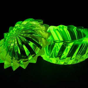 Könnte beinhalten: Eine leuchtend grüne Glas-Schmuckschatulle mit Deckel, vor schwarzem Hintergrund. Die Schatulle und der Deckel haben ein radiales, geripptes Design, das einen Sternausbruch-Effekt erzeugt. Die offene Schatulle zeigt ihr Inneres.