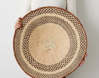 Pode incluir: Uma cesta de parede redonda e trançada, com um centro em tom bege natural e um detalhe quadrado. A cesta apresenta uma borda larga com um padrão geométrico repetitivo em castanho escuro e bege, e um aro espesso em castanho escuro.